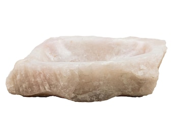 Puede incluir: Un cuenco de sal de roca rosa claro, natural, toscamente tallado. El cuenco está tallado en una sola pieza de roca y tiene una forma natural y orgánica.
