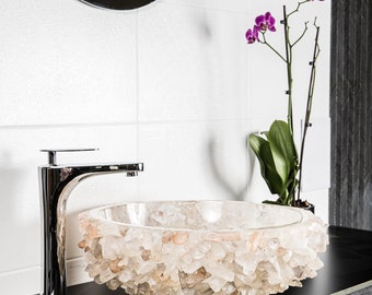 May include: A white crystal quartz sink with a chrome faucet. The sink is on a black countertop with a drawer. A pink orchid is in a pot on the counter.