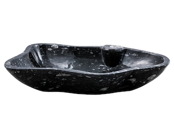 May include: A black fossilized stone sink with a unique, organic shape. The sink has a smooth, polished surface and features a drain hole in the center.