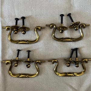 May include: Four sets of brass-toned drawer pulls with black screws. Each pull has a curved handle and two screw posts. The pulls are arranged on a neutral-colored fabric background. These are suitable for furniture restoration or DIY projects.