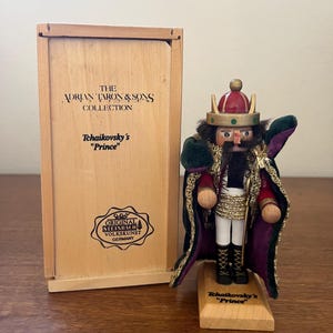 May include: A wooden nutcracker figurine of a prince, wearing a red and gold crown, a red and purple cape, and black boots. The figurine is on a wooden base that reads "Tchaikovsky's 'Prince'". The wooden box next to it reads "The Adrian Taron & Sons Collection".