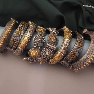 Könnte beinhalten: Eine Sammlung von silber- und goldfarbenen Armreifen. Die Armreifen weisen aufwendige Designs auf, darunter Blumenmuster, geometrische Formen und Perlenakzente. Der Schmuck wird auf einer blauen Oberfläche vor einem dunkelgrünen Hintergrund präsentiert.