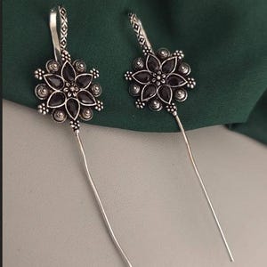 Puede incluir: Un par de pendientes plateados con diseño floral. Cada pendiente presenta un grupo de gemas negras en forma de flor, rodeado por un marco plateado detallado. Largos y delgados alambres plateados se extienden desde el diseño floral.