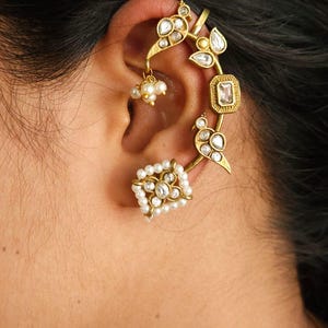 May include: Gold-toned ear cuff featuring a floral and bird design, adorned with clear gemstones and small white pearls. The cuff wraps around the ear, with a square stud earring at the bottom. A cluster of pearls hangs from the design.