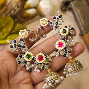 May include: Ornate silver and gold-toned earrings with colorful gemstones and faux pearls. The earrings feature a floral design with blue, green, and pink stones, and a bell-shaped dangle. The earrings are held in a hand.