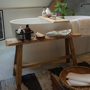 Könnte beinhalten: Eine weiße Badewanne mit einem Holztablett, auf dem Flaschen und ein Glas stehen. Die Badewanne befindet sich in einem Badezimmer mit einer Holzbalkendecke und einem grauen Fliesenboden. Es gibt einen Holzhocker mit einem Korb und einem Magazin darauf. Ein geflochtener Korb mit Handtüchern steht auf dem Boden.