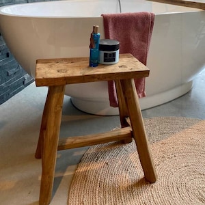 Peut inclure: Un tabouret en bois avec une finition naturelle se trouve dans une salle de bain avec une baignoire ovale blanche, une serviette rose et un tapis en jute. Deux flacons bleus et un pot de crème sont sur le tabouret.