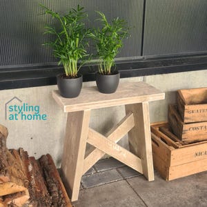 Op de afbeelding: Een lichtgekleurde houten kruk met een X-vormig onderstel, waarop twee groene kamerplanten in donkergrijze potten staan. De tekst "styling at home" is links zichtbaar. De kruk staat in de buurt van houten kratten.