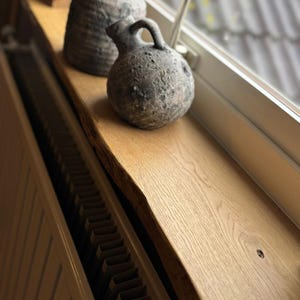 May include: A light oak wooden windowsill with a live edge, featuring two textured, gray ceramic decorative pieces. The window frame is white, and a radiator is visible below the sill. The objects are illuminated by natural light.