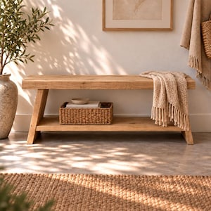 Op de afbeelding: Een rechthoekige lichtbruine houten bank met een onderste plank en schuine poten. Op de onderste plank staat een gevlochten rechthoekige mand en een beige, getextureerde deken met franjes hangt over de rechterkant van de zitting van de bank.