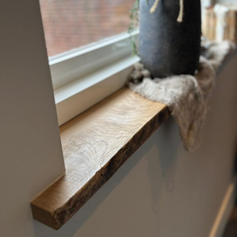 May include: A natural wood window sill with a live edge. The light-colored wood has a visible grain and is installed against a white wall. A dark gray pot and a piece of fabric sit on the sill near the window.