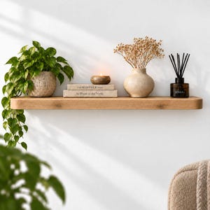 Peut inclure: Une étagère flottante en bois clair supportant une plante verte en pot, deux livres empilés intitulés The Kinfolk Home et At Home in the North, une petite bougie ronde, un vase beige avec des fleurs séchées et un diffuseur en verre foncé.