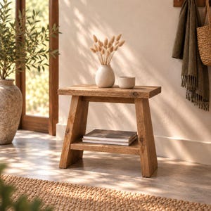 Op de afbeelding: Een kleine houten bijzettafel met een rechthoekig blad en een onderste plank. De tafel heeft schuine poten en een natuurlijke houtnerfafwerking. Op het blad staan een lichtgekleurde keramische vaas met gedroogde grassen en een kommetje.