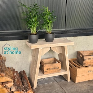 Peut inclure: Une table d'appoint en bois clair avec une étagère inférieure contenant une caisse en bois marquée GUATEMALA. Deux plantes vertes dans des pots gris foncé sont posées sur le dessus. Sur le côté, du bois de chauffage et une caisse sont visibles.