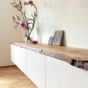 Könnte beinhalten: Ein schwebender weißer Schrank mit einer natürlichen Holzplatte. Das Holz hat eine rohe Kante und eine hellbraune Farbe. Eine Glasvase mit rosa Blüten und eine kleine rosa Lotusblume stehen oben. Zwei kleine dekorative Fliesen sind ebenfalls vorhanden.