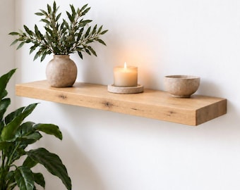 May include: A light brown wooden floating shelf mounted on a wall. On the shelf sits a round beige vase containing a green leafy plant, a lit candle in a beige holder, and a small beige bowl. A large green leafy plant is visible in the foreground.