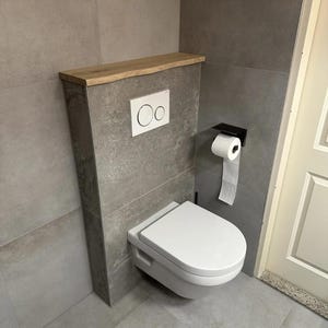 May include: A modern, wall-mounted toilet with a white seat and lid. The toilet is set against a gray tiled wall, with a wooden shelf above. A black toilet paper holder and a white flush button are also visible.