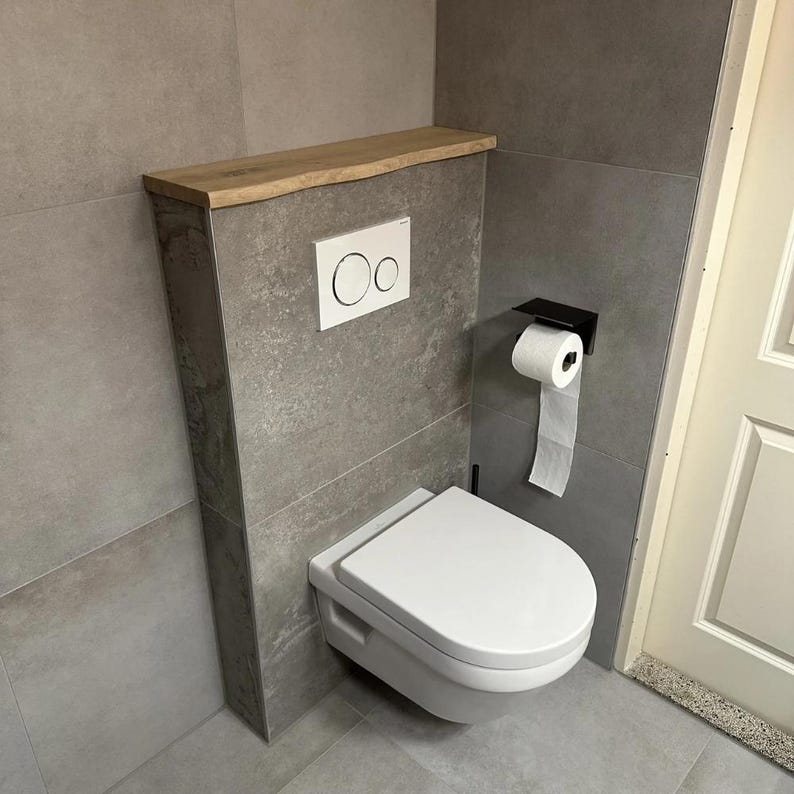 May include: A modern, wall-mounted toilet with a white seat and lid. The toilet is set against a gray tiled wall, with a wooden shelf above. A black toilet paper holder and a white flush button are also visible.
