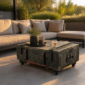 Op de afbeelding: Een verweerde, donkergroene salontafel gemaakt van een hergebruikte militaire kist. De tafel heeft metalen hardware, wielen en een rode streep. Een kleine plant, een kaars en een glas amberkleurige vloeistof staan erop.