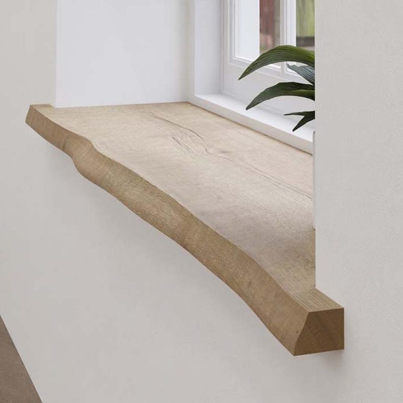 May include: A natural wood window sill with a live edge. The light brown sill is mounted on a white wall, with a window and greenery visible in the background. The wood grain is visible on the surface.