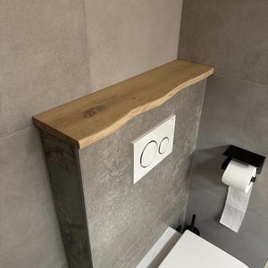May include: A wooden shelf with a live edge is mounted above a toilet. The shelf is light brown and has a natural, organic shape. The wall behind the toilet is gray, and a white flush button and a black toilet paper holder are visible.