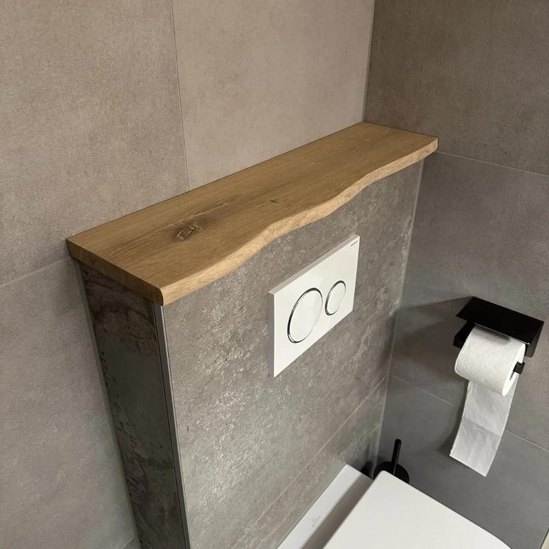 May include: A wooden shelf with a live edge is mounted above a toilet. The shelf is light brown and has a natural, organic shape. The wall behind the toilet is gray, and a white flush button and a black toilet paper holder are visible.
