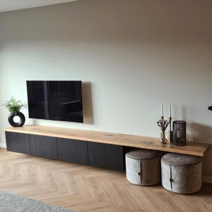 Könnte beinhalten: Ein modernes Unterhaltungscenter mit schwarzem Schrankfuß und heller Holzplatte. Ein schwarzer Fernseher ist über dem Schrank montiert. Zwei grau gepolsterte Ottomanen stehen vor dem Schrank. Eine schwarze Vase und Kerzen stehen oben.