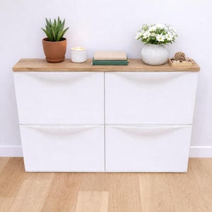 Könnte beinhalten: Weißer Schrank mit vier Schubladen und einer hellen Holzplatte. Eine Topfpflanze, eine Kerze, Bücher und eine Vase mit weißen Blumen schmücken die Oberseite. Der Schrank steht auf einem hellen Holzboden.