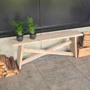 Op de afbeelding: Een lichtgekleurde houten bank met een platte rechthoekige zitting en een onderstel met twee schuine poten verbonden door een diagonale steunbalk. Twee kleine groene kamerplanten in donkergrijze potten staan links op de zitting van de bank.