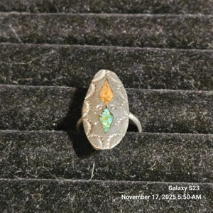 Puede incluir: Anillo ovalado de plata con incrustaciones de piedras naranja y turquesa. El anillo tiene un borde decorativo y está sobre un fondo negro. El anillo es de talla 14.