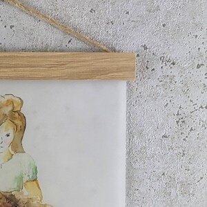 Puede incluir: Una pintura de acuarela de una ni&ntilde;a con cabello largo y casta&ntilde;o que sostiene un perro marr&oacute;n y blanco. La pintura est&aacute; enmarcada con un marco de madera marr&oacute;n claro y colgada de una cuerda.