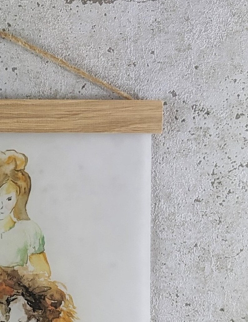 Puede incluir: Una pintura de acuarela de una ni&ntilde;a con cabello largo y casta&ntilde;o que sostiene un perro marr&oacute;n y blanco. La pintura est&aacute; enmarcada con un marco de madera marr&oacute;n claro y colgada de una cuerda.