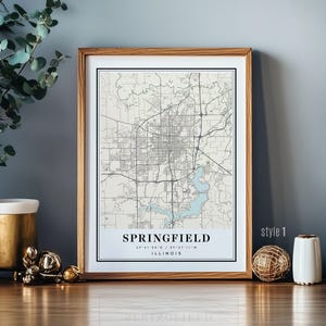 Puede incluir: Impresión enmarcada de un mapa de Springfield, Illinois, en un marco de madera clara. El mapa presenta un diseño detallado de las calles, con un lago azul claro y el nombre de la ciudad en la parte inferior. El texto "style 1" está en la esquina inferior derecha.