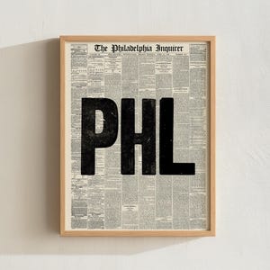 May include: A framed print featuring a page from The Philadelphia Inquirer newspaper with the large, bold, black letters PHL printed over the center of the text columns. The frame is made of light-colored wood and has a simple, clean design.