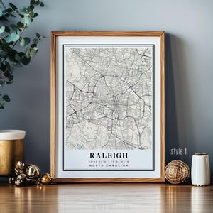 May include: A framed map print of Raleigh, North Carolina, in a minimalist style. The map shows a network of roads and streets in black and white, with the city name and coordinates at the bottom. The frame is made of light brown wood.