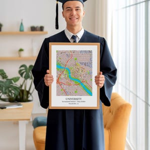 May include: A framed map print featuring a detailed city plan with a river running through it. The print is held by a person wearing a graduation gown and cap. The text on the print reads "UNIVERSITY Environmental Science - Class of 2024 PHILADELPHIA, GA."