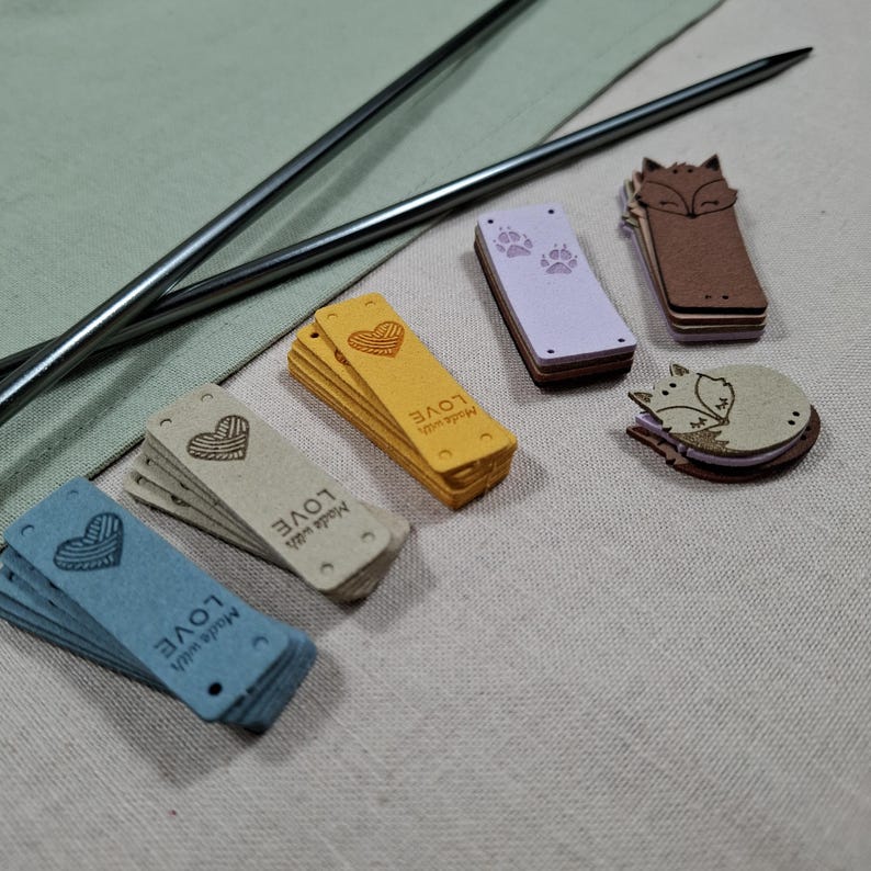 Op de afbeelding: Een verzameling knutselbenodigdheden, waaronder breinaalden, decoratieve labels en houten versieringen. De labels zijn in verschillende kleuren, met hart- en pootafdrukontwerpen, en de woorden "Made with LOVE". Houten vossen vormen zijn ook aanwezig.