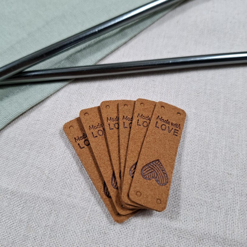 Op de afbeelding: Een waaier van bruine, rechthoekige labels met afgeronde hoeken, elk bedrukt met "Made with LOVE" en een hartmotief. De labels liggen op een lichte stof, met op de achtergrond twee zilveren metalen rietjes.