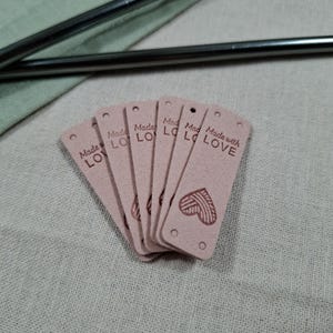 Op de afbeelding: Een stapel roze rechthoekige labels met afgeronde hoeken en kleine gaatjes aan de bovenkant. Elk label heeft de tekst "Made with LOVE" en een hartontwerp. De labels zijn gemaakt van een zacht materiaal.