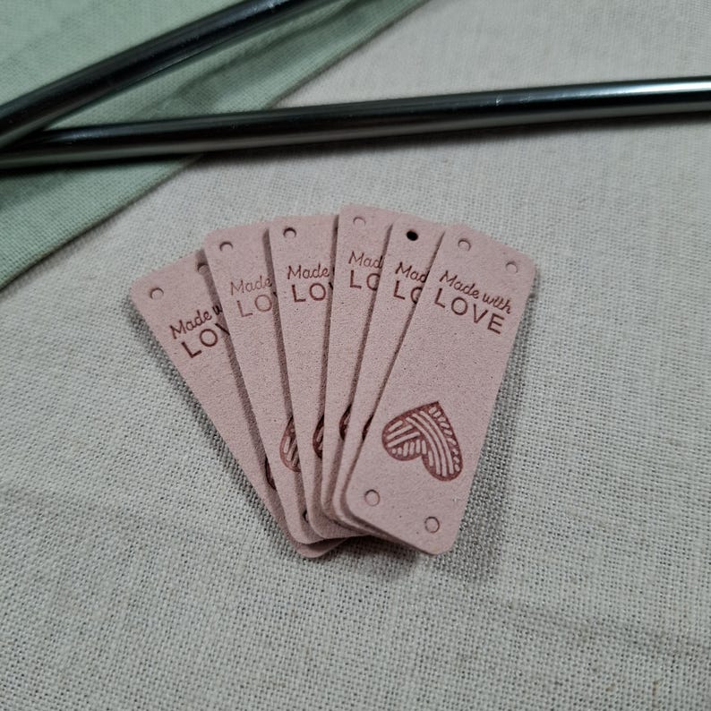 Op de afbeelding: Een stapel roze rechthoekige labels met afgeronde hoeken en kleine gaatjes aan de bovenkant. Elk label heeft de tekst "Made with LOVE" en een hartontwerp. De labels zijn gemaakt van een zacht materiaal.