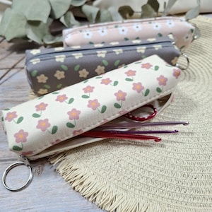 Könnte beinhalten: Drei rechteckige Stiftemäppchen aus Stoff mit Reißverschluss und Metallring. Das vordere Etui ist cremefarben mit rosa Blumen, das mittlere grau mit gelben Blumen und das hintere hellrosa mit weißen Blumen. Im Inneren sind Häkelnadeln zu sehen.