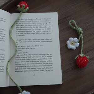 May include: Crocheted strawberry and flower bookmarks. The strawberry bookmark is red with green leaves, and the flower bookmark is white with a yellow center. The bookmarks are on a book.