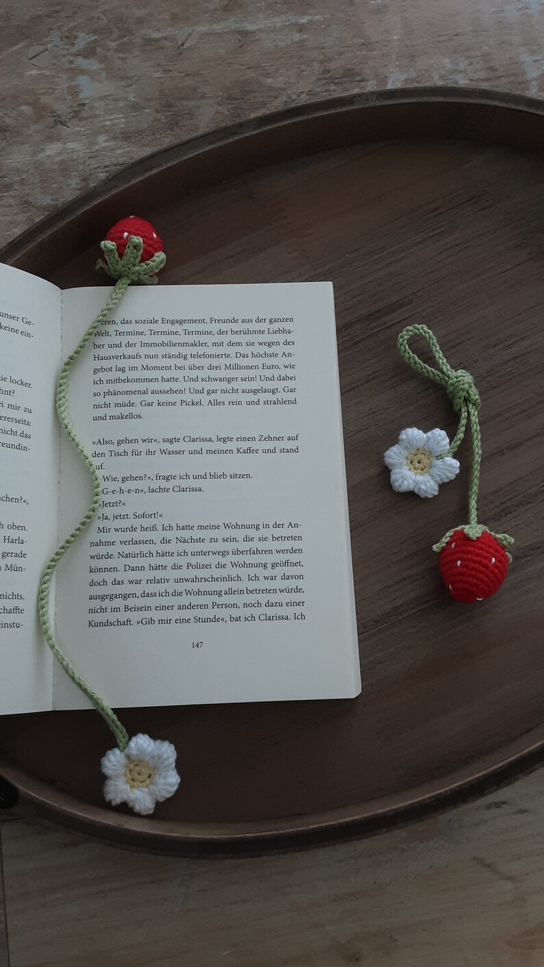 May include: Crocheted strawberry and flower bookmarks. The strawberry bookmark is red with green leaves, and the flower bookmark is white with a yellow center. The bookmarks are on a book.