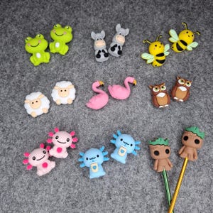 Könnte beinhalten: Eine Sammlung kleiner, bunter Silikonanhänger mit verschiedenen Figuren, darunter grüne Frösche, graue Esel, gelbe Bienen, weiße Schafe, rosa Flamingos, braune Eulen, rosa und blaue Axolotls sowie zwei braune baumartige Figuren an Metallstäben.