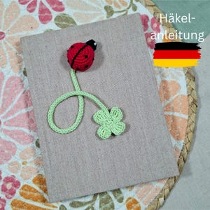 Könnte beinhalten: Handgemachter Lesezeichen mit einem rot-schwarzen Marienkäfer mit grünem Stiel und vierblättrigem Kleeblatt. Das Lesezeichen liegt auf einem beigen Buch. Das Bild enthält auch den Text "Häkelanleitung" und die deutsche Flagge.