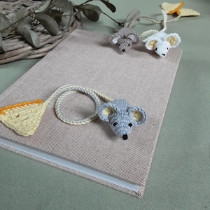 Puede incluir: Tres marcapáginas de ratón de ganchillo con detalles de queso. Los ratones son grises y blancos y el queso es amarillo. Los marcapáginas están en una libreta marrón.