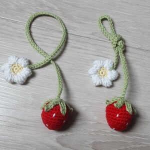 May include: Two handmade crochet strawberry ornaments. Each ornament features a red strawberry with white seed details, green leaves, and a green stem. A white daisy with a yellow center is attached to the stem. The ornaments are connected by a green cord.