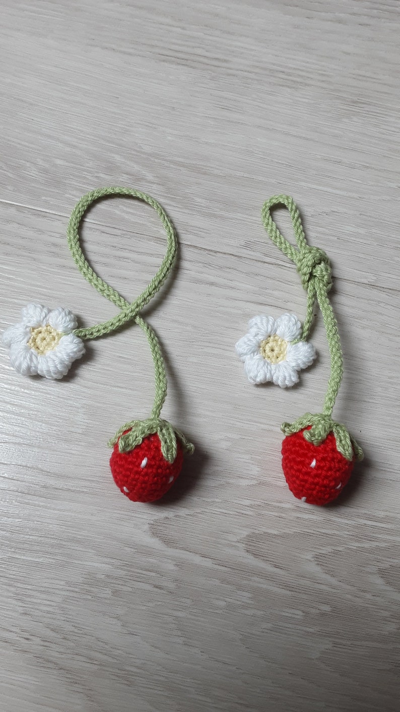 May include: Two handmade crochet strawberry ornaments. Each ornament features a red strawberry with white seed details, green leaves, and a green stem. A white daisy with a yellow center is attached to the stem. The ornaments are connected by a green cord.