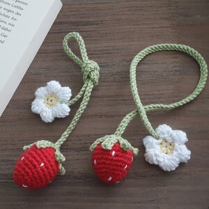 May include: Two handmade crochet strawberry bookmarks with white flower accents. The strawberries are red with white seed details and green tops. The bookmarks are on a wooden surface next to a book.