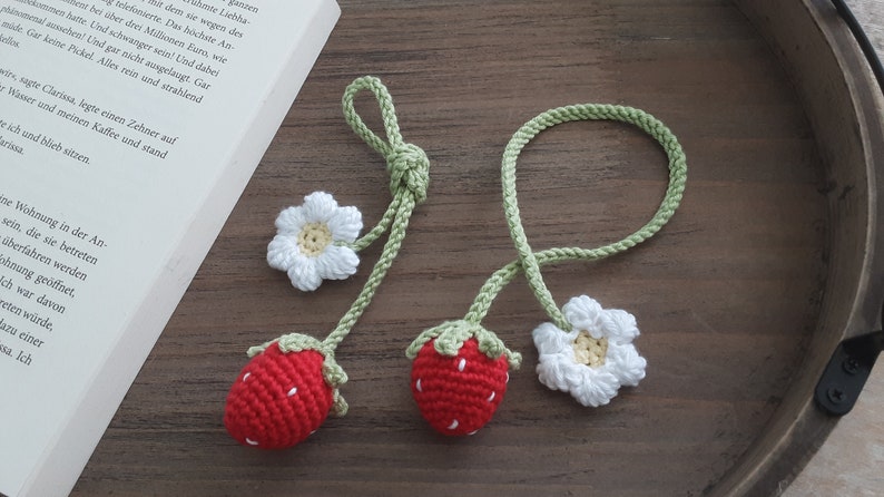 May include: Two handmade crochet strawberry bookmarks with white flower accents. The strawberries are red with white seed details and green tops. The bookmarks are on a wooden surface next to a book.
